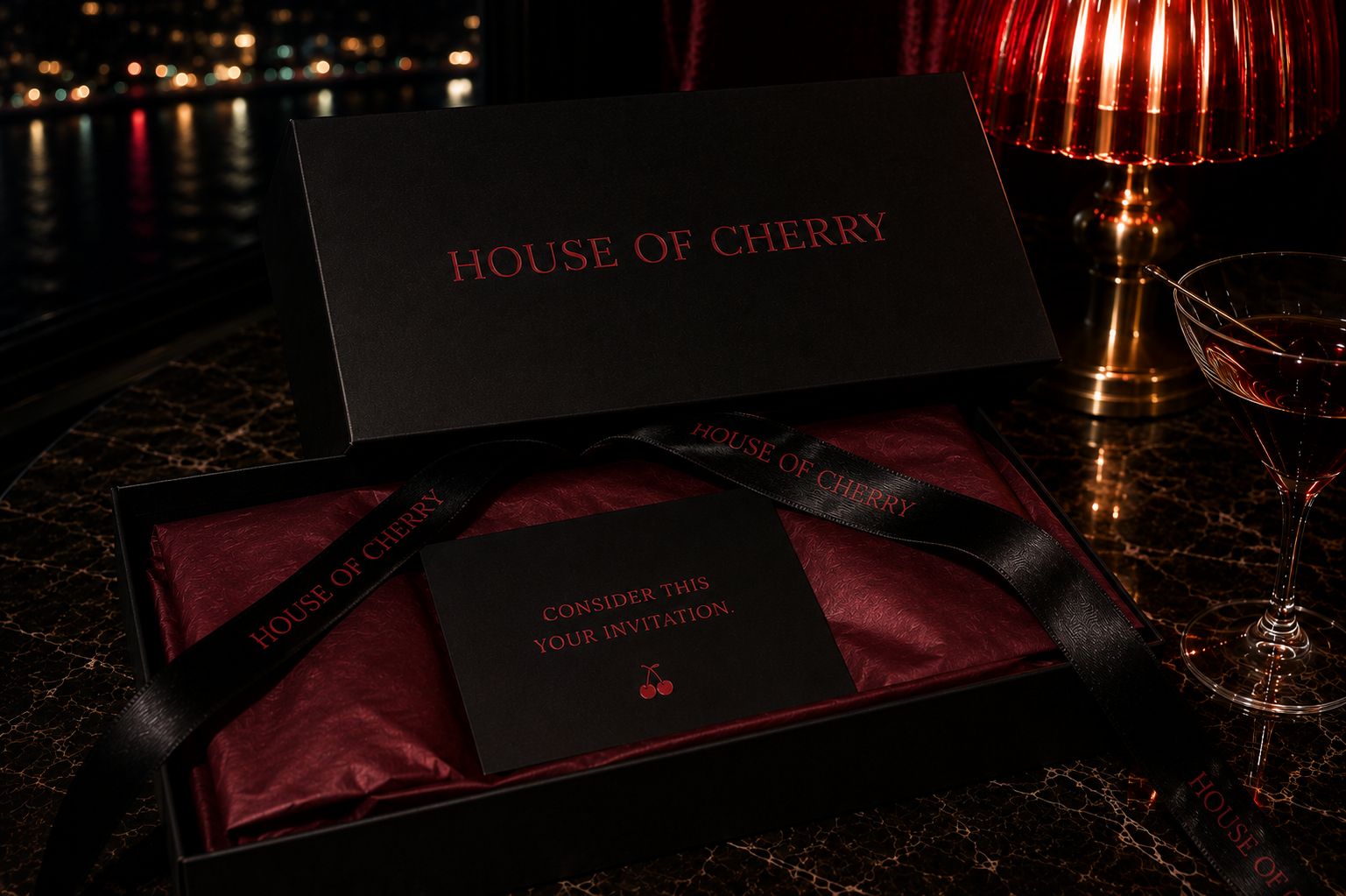 A luxury gift box set from House of Cherry, featuring a black lid with gold lettering, a black ribbon, and a black card with gold text, presented on a dark marble table next to a red lamp and a glass of red wine.